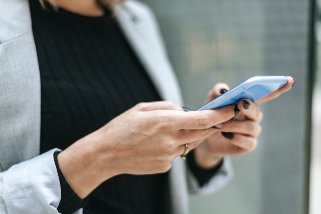 Imagem de uma mulher com uma blusa preta e blazer cinza, segurando um celular com as duas mãos