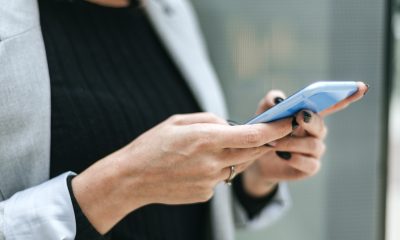 Imagem de uma mulher com uma blusa preta e blazer cinza, segurando um celular com as duas mãos