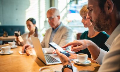 Imagem de pessoas em volta de uma mesa redonda de trabalho, utilizando celular, notebook e outros meios de trabalho